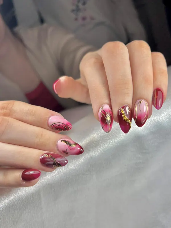 Uñas almendradas en burdeos y rosa con líneas doradas y bolitas metálicas